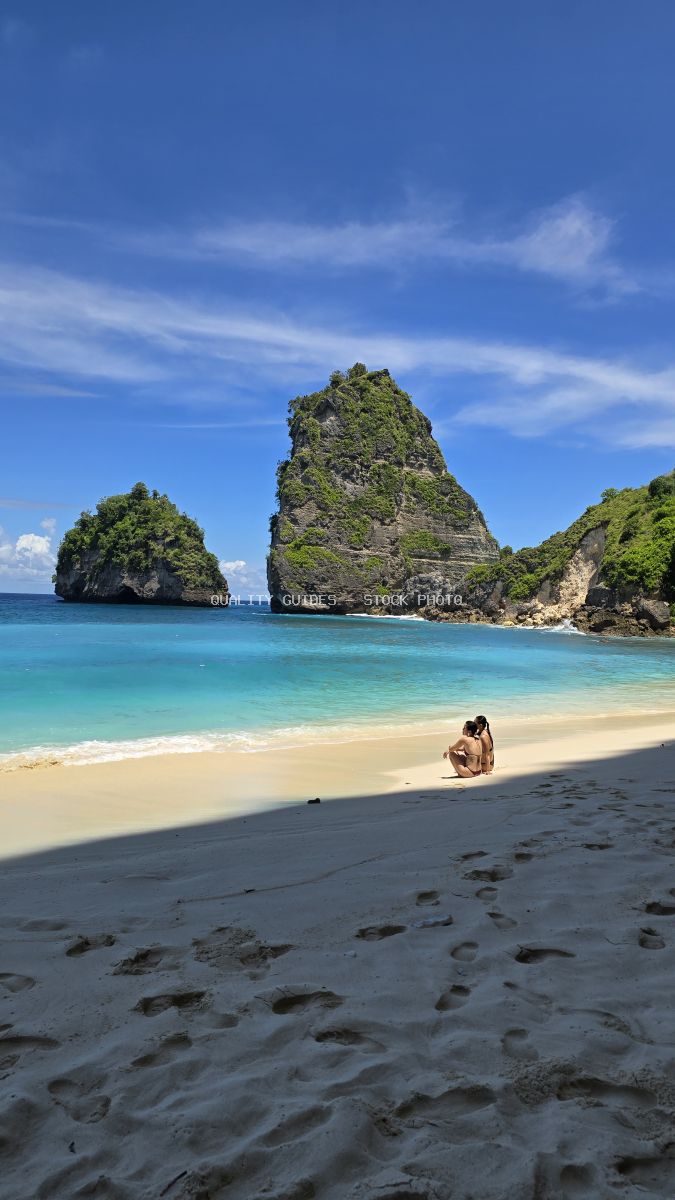 Person sitting on white sand beach facing turquoise ocean with dramatic green-covered rock formations at Diamond Beach Bali - stock photo