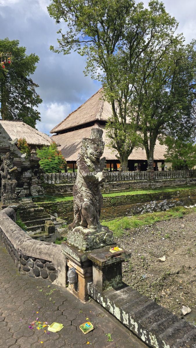 Stone carved statue at Taman Ayun Temple in Bali with thatched roof temple building and green trees - stock photo