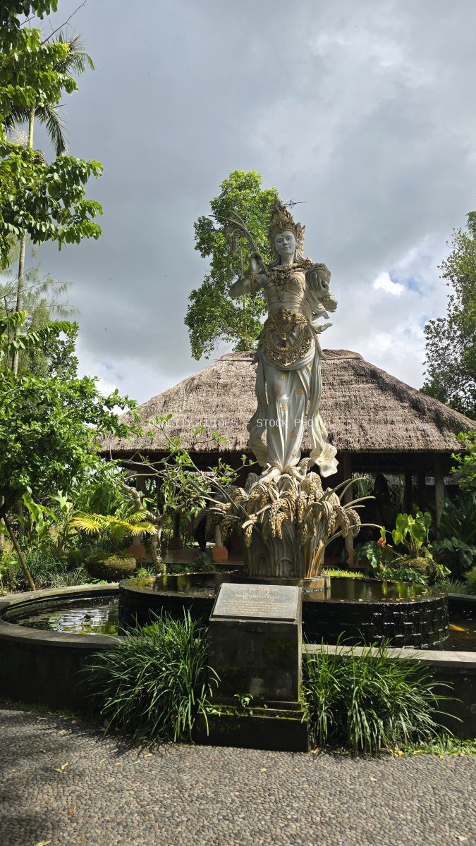 Stone deity statue covered in green vines at Taman Ayun Temple courtyard in Bali, with traditional thatched-roof building in background - stock photo