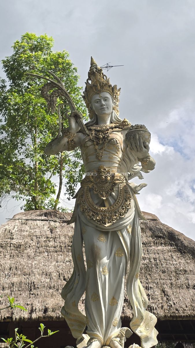 Ornate stone goddess statue with crown and ceremonial staff at Taman Ayun Temple in Bali, with traditional thatched-roof structure and green foliage in background - stock photo