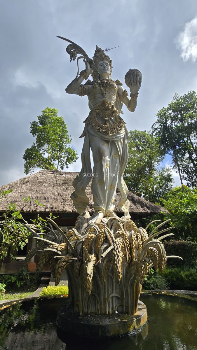 Bronze Hindu goddess statue with multiple arms on lotus pedestal in pond at Taman Ayun Temple, Bali, with traditional thatched structures and tropical trees in background - stock photo