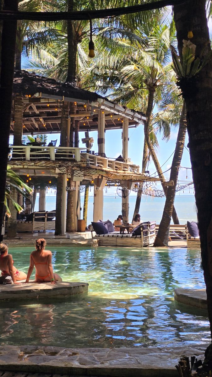 Tropical beach club with wooden structure, turquoise pool, palm trees, and swimmers in Bali - stock photo