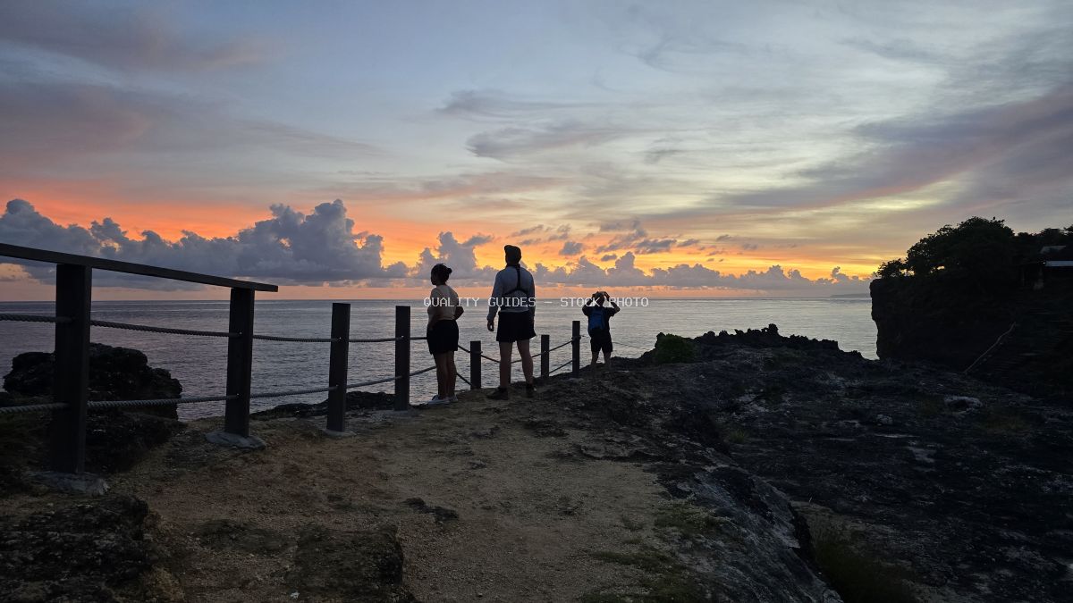 Silhouetted tourists watching sunset at Broken Beach Bali with dramatic orange sky and wooden railing - stock photo