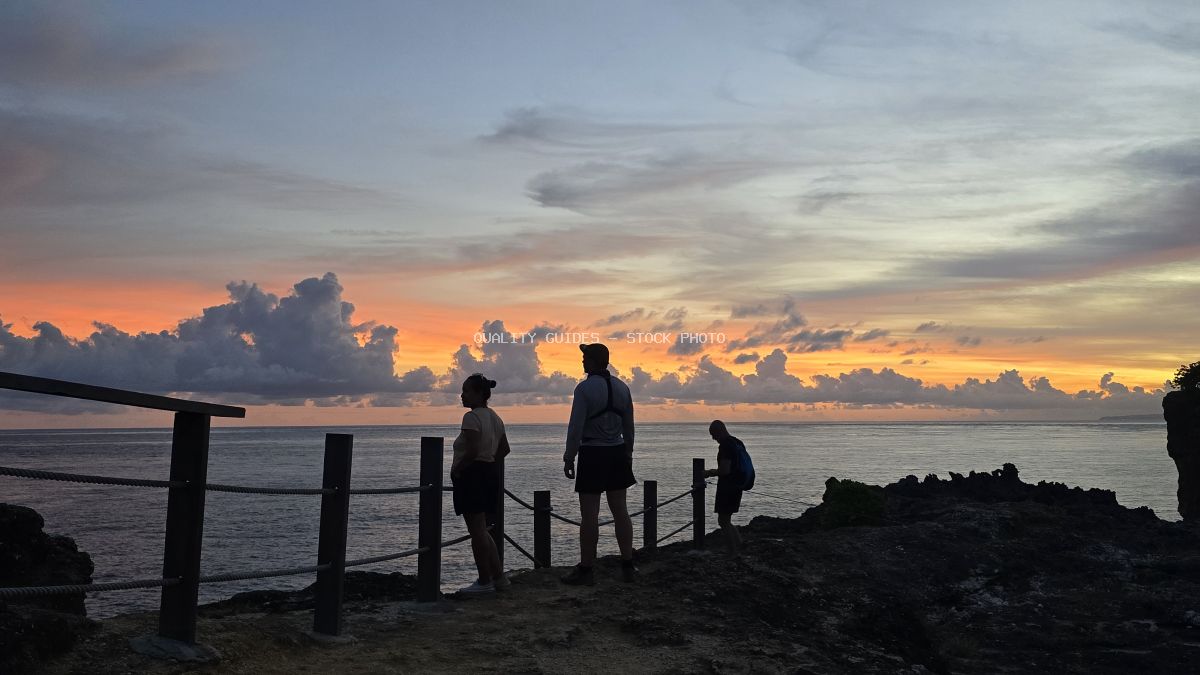 Silhouetted tourists viewing sunset at Broken Beach Bali with wooden railings and dramatic sky - stock photo