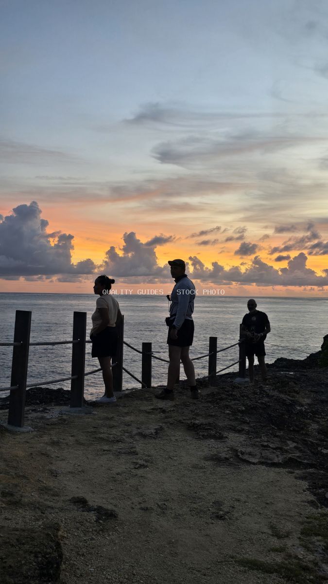 Silhouetted tourists watching golden sunset at Broken Beach in Bali with wooden barrier and calm ocean - stock photo