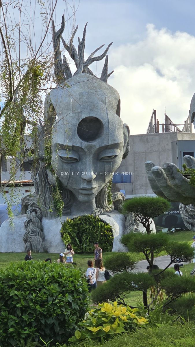 Surreal stone head sculpture with reaching hands at Nuanu Creative City in Bali, surrounded by tropical gardens and visitors - stock photo