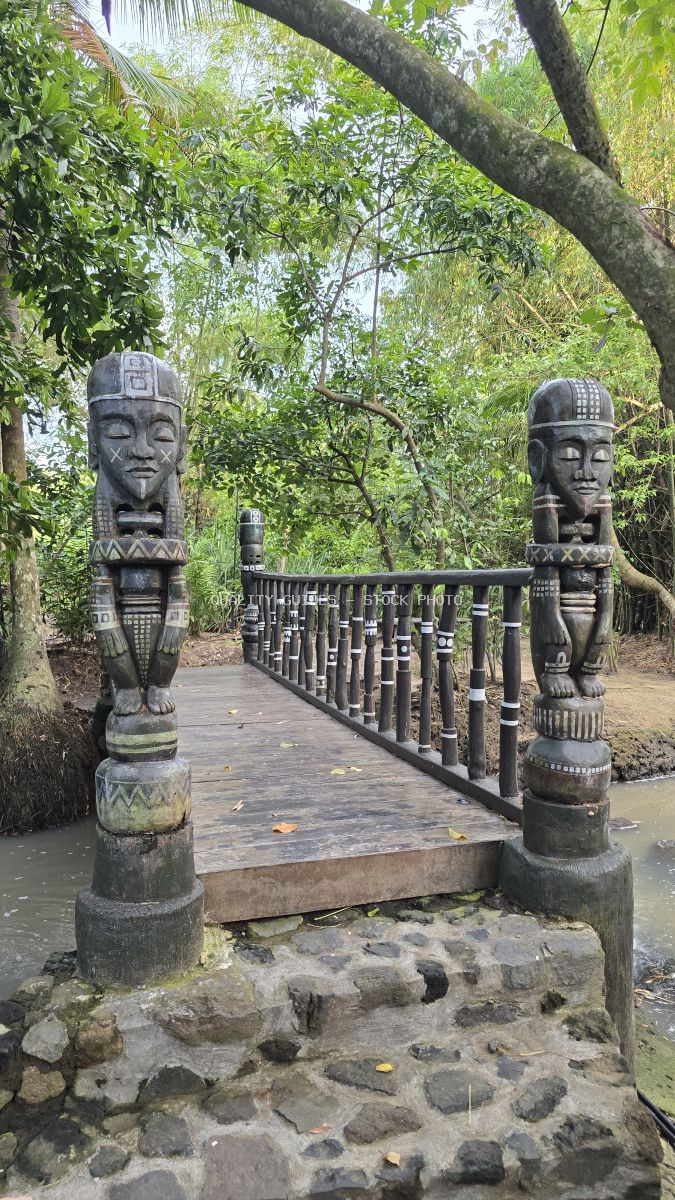 Two carved Balinese stone statues flanking a wooden bridge in a lush tropical garden with green foliage overhead - stock photo