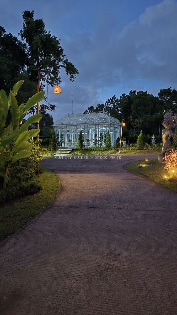 Illuminated glass greenhouse at Nuanu Creative City in Bali at dusk, surrounded by tropical gardens and curved brick pathway - stock photo