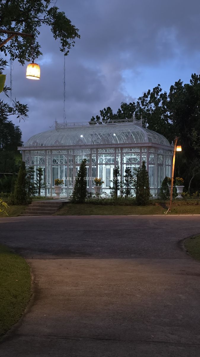 Illuminated white glass greenhouse with domed roof at twilight, surrounded by tropical plants and hanging lights in Bali - stock photo