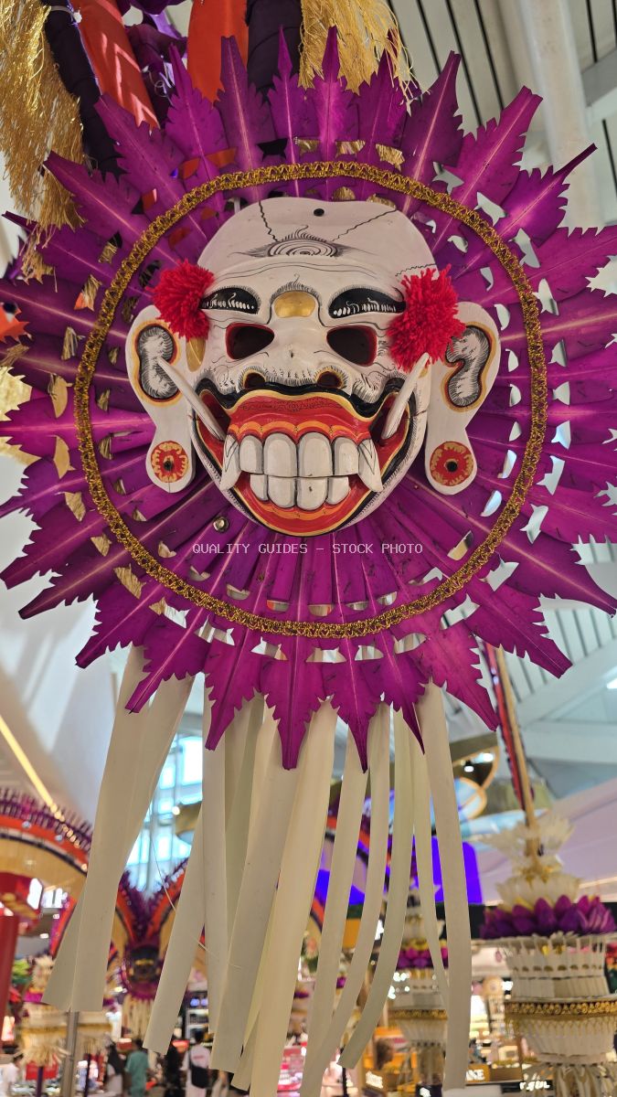 Traditional purple and gold Balinese demon mask decoration hanging in Bali airport terminal with feathered crown and grinning expression - stock photo