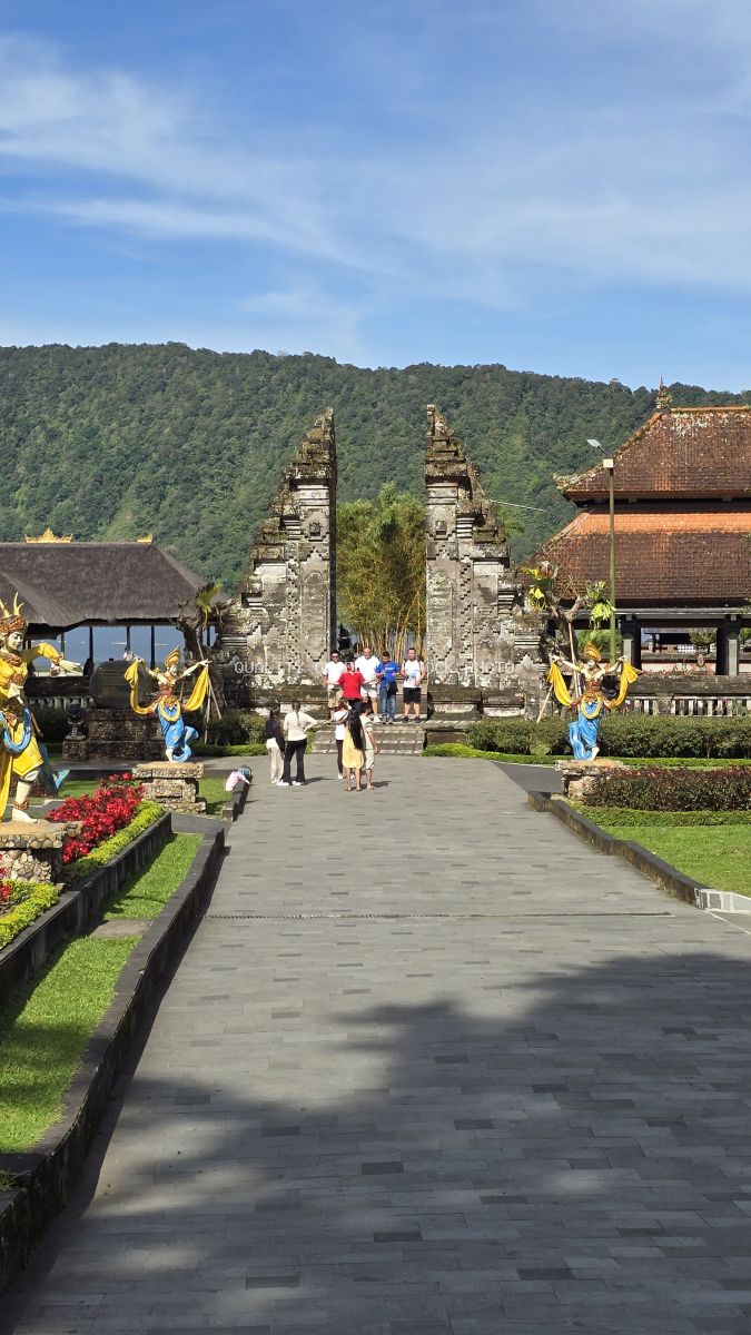 Tourists walking through the ornate stone gateway entrance of Ulun Danu Beratan Temple in Bali with lush green mountains in background - stock photo