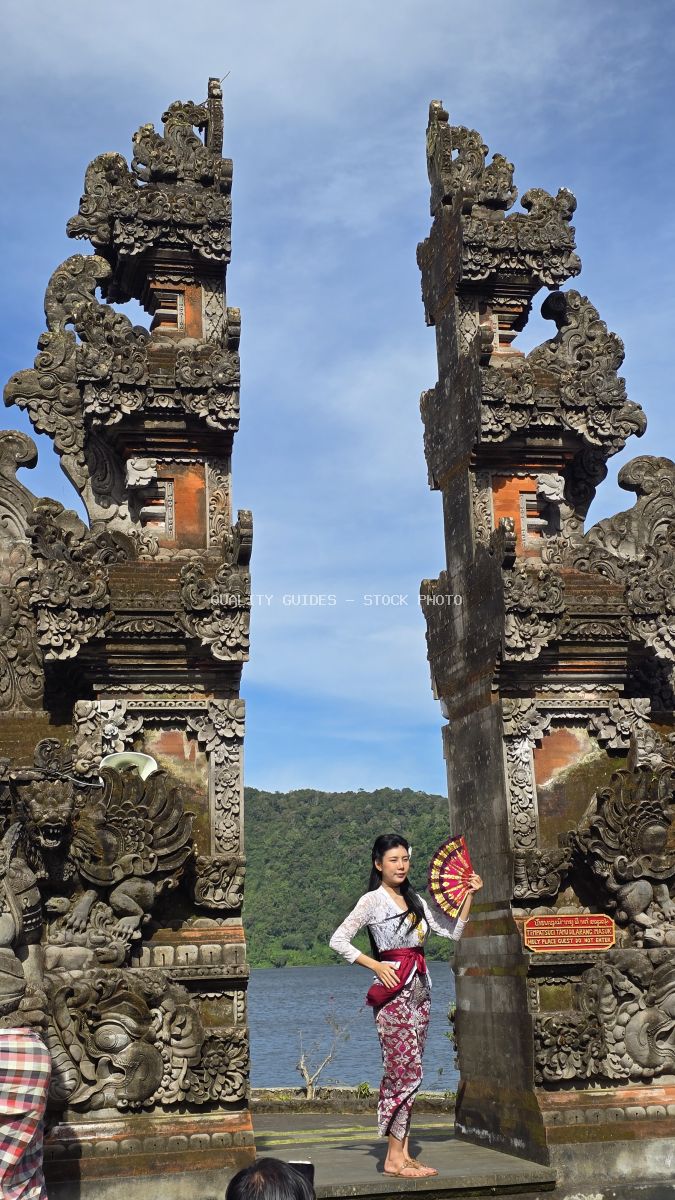 Woman in traditional Balinese dress posing between ornate carved stone gates at Ulun Danu Beratan Temple with lake and mountains in background - stock photo