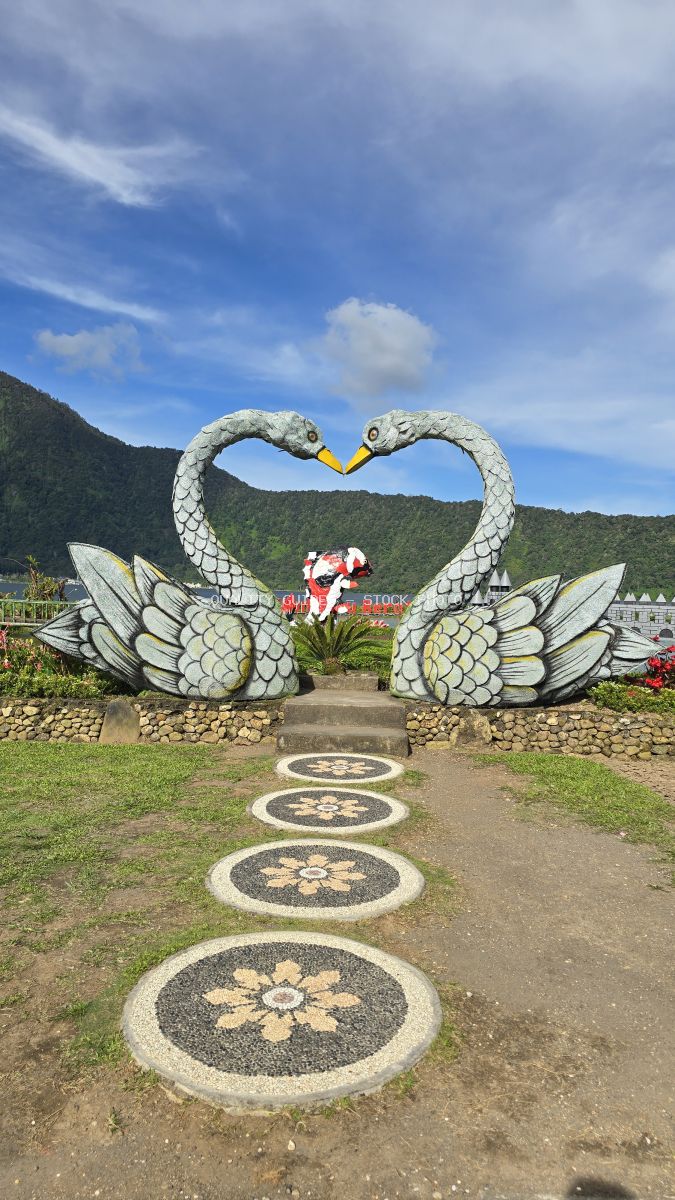 Two white decorative swans forming a heart shape in a Bali temple garden with mountains and blue sky - stock photo