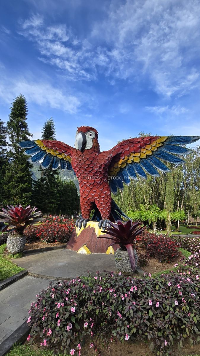Large red and blue macaw statue in tropical Bali garden with pink flowers and blue sky - stock photo