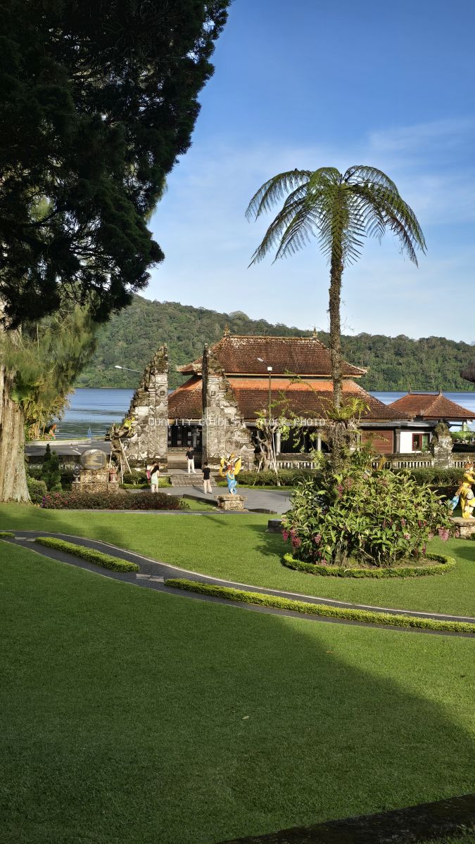 Ulun Danu Beratan Temple in Bali with palm tree, lake, and manicured gardens under blue sky - stock photo