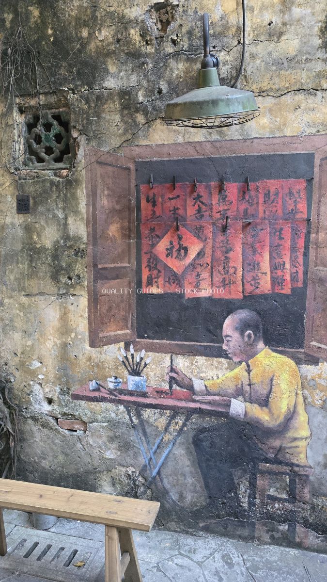 Street art mural of man in yellow shirt doing calligraphy with red Chinese characters and vintage lamp on weathered wall in Kuala Lumpur - stock photo