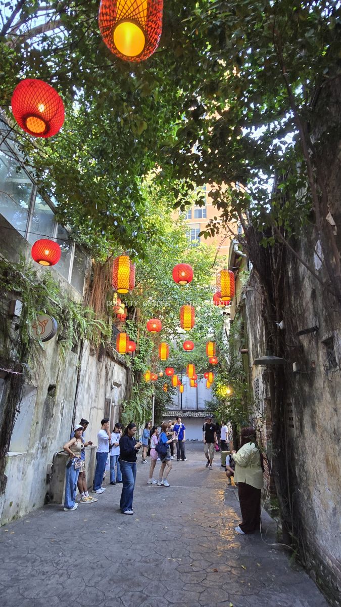 Vibrant red and yellow lanterns hanging in a tree-lined historic alley with tourists walking through - stock photo